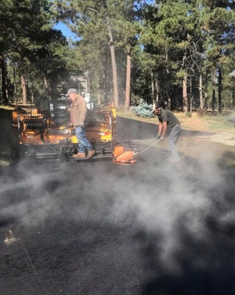 Dawley Asphalt crew actively installing 3.5 inch hot mix asphalt driveway with steam rising from fresh material through pine lined residential property in Monument Colorado