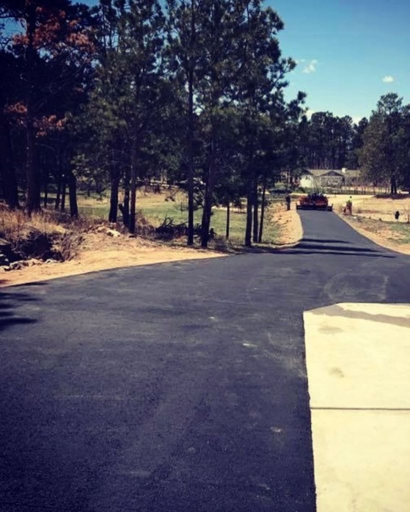 Newly installed 3.5 inch hot mix asphalt driveway with compaction roller at far end and concrete apron street connection through pine lined Monument Colorado residential property by Dawley Asphalt