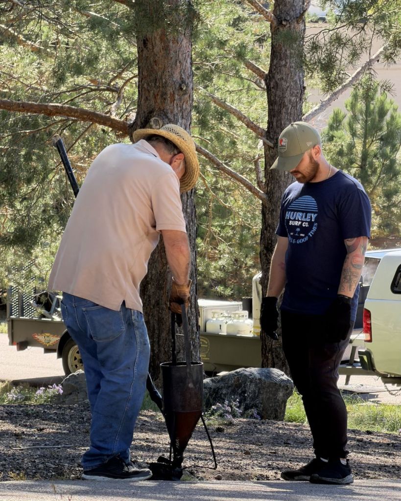 Jerry Dawley's father, James Dawley, and crew member applying hot rubberized crack fill on residential driveway in Winsome neighborhood Colorado Springs with pine trees and trailer in background