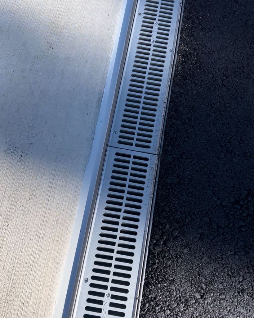 Custom stainless steel linear trench drain installed flush between concrete garage apron and asphalt driveway at sloped Woodmoor residence in Monument Colorado by Dawley Asphalt